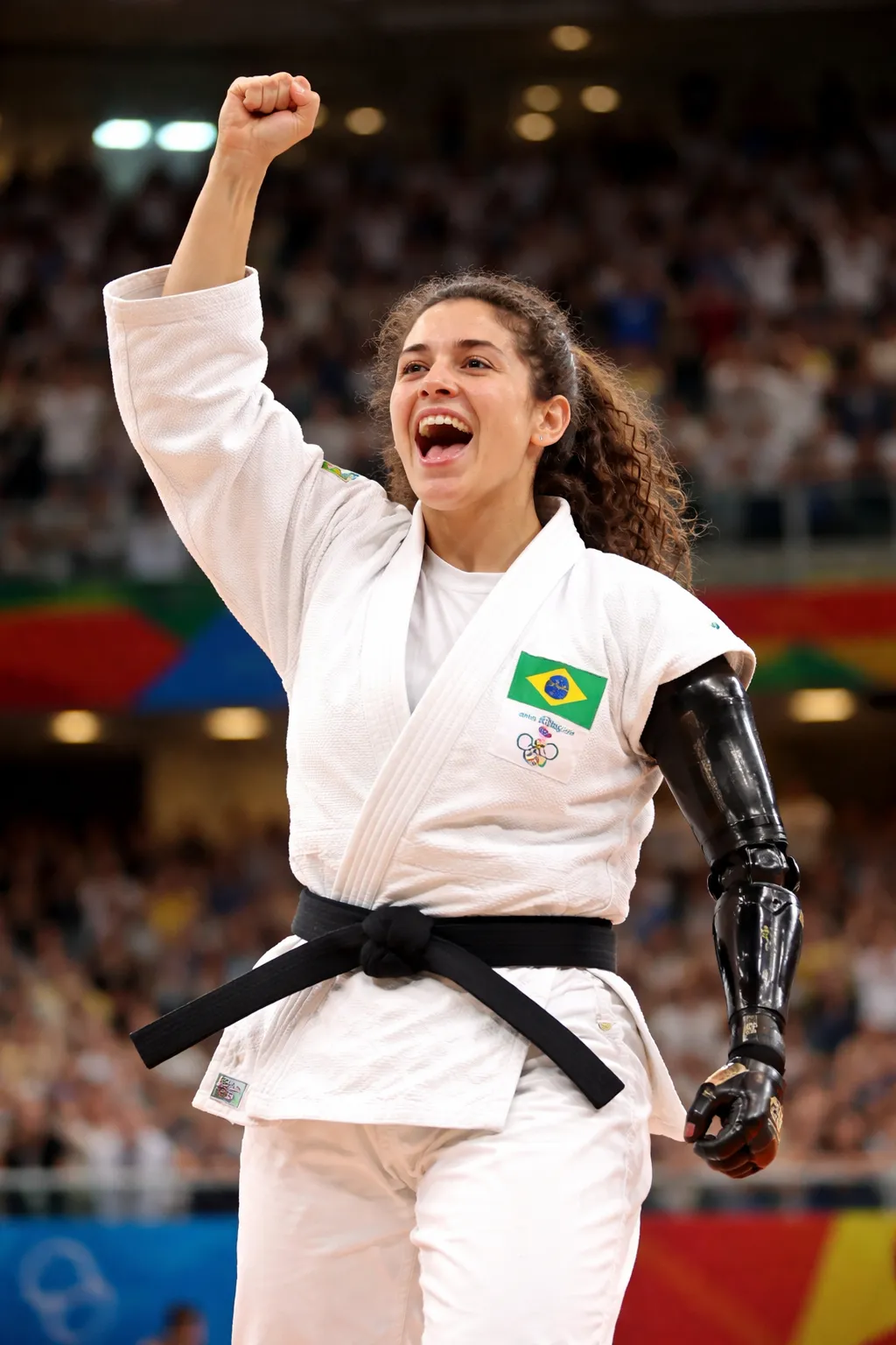 Uma atleta de judô com quimono branco e faixa preta, comemorando com o punho esquerdo erguido e a boca aberta em expressão de alegria. O braço direito é uma prótese mecânica preta, brilhante, com articulações visíveis. No peito do quimono há um patch com a bandeira do Brasil e, abaixo, pequenos símbolos dos anéis olímpicos; o texto é pequeno demais para leitura nítida. O cabelo é castanho e cacheado, preso para trás, e a pele é clara. Ao fundo há uma arena lotada e desfocada, com luzes fortes no alto.