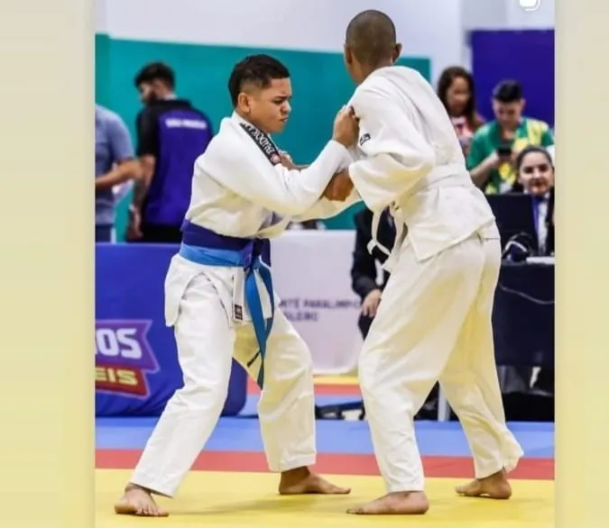 Dois praticantes de judô em um tatame, durante uma luta. Ambos usam quimono branco; o atleta da esquerda é Michel e tem faixa azul e está com os pés afastados, segurando a gola e a manga do adversário, com expressão concentrada. O atleta da direita está de costas para a câmera, também segurando o quimono do oponente, e usa faixa branca. Ao fundo há pessoas em um ginásio, incluindo alguém sentado em uma mesa com um laptop, e banners/placas parcialmente desfocados.
