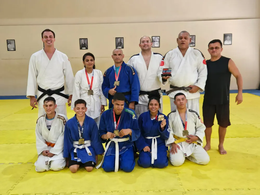 Um grupo de nove pessoas posando em um tatame amarelo, em um dojo, usando quimonos de judô (brancos e azuis) e exibindo medalhas.
Na fileira de trás há cinco pessoas em pé. Três homens de quimono branco (um bem alto com faixa preta; outro de estatura média com faixa preta; e um homem mais alto e mais largo com faixa preta segurando um troféu/placa). Entre eles há um homem de quimono azul com medalhas no pescoço. Ao lado esquerdo, há uma mulher de quimono branco com medalha. À extrema direita, há um homem sem quimono, usando camiseta preta sem mangas e bermuda escura.
Na fileira da frente há quatro pessoas ajoelhadas. Um homem de quimono branco à esquerda, com medalha e faixa laranja. Três pessoas de quimono azul e uma de quimono branco à direita (com faixa amarela), todas com medalhas; uma das pessoas na frente segura uma medalha voltada para a câmera.